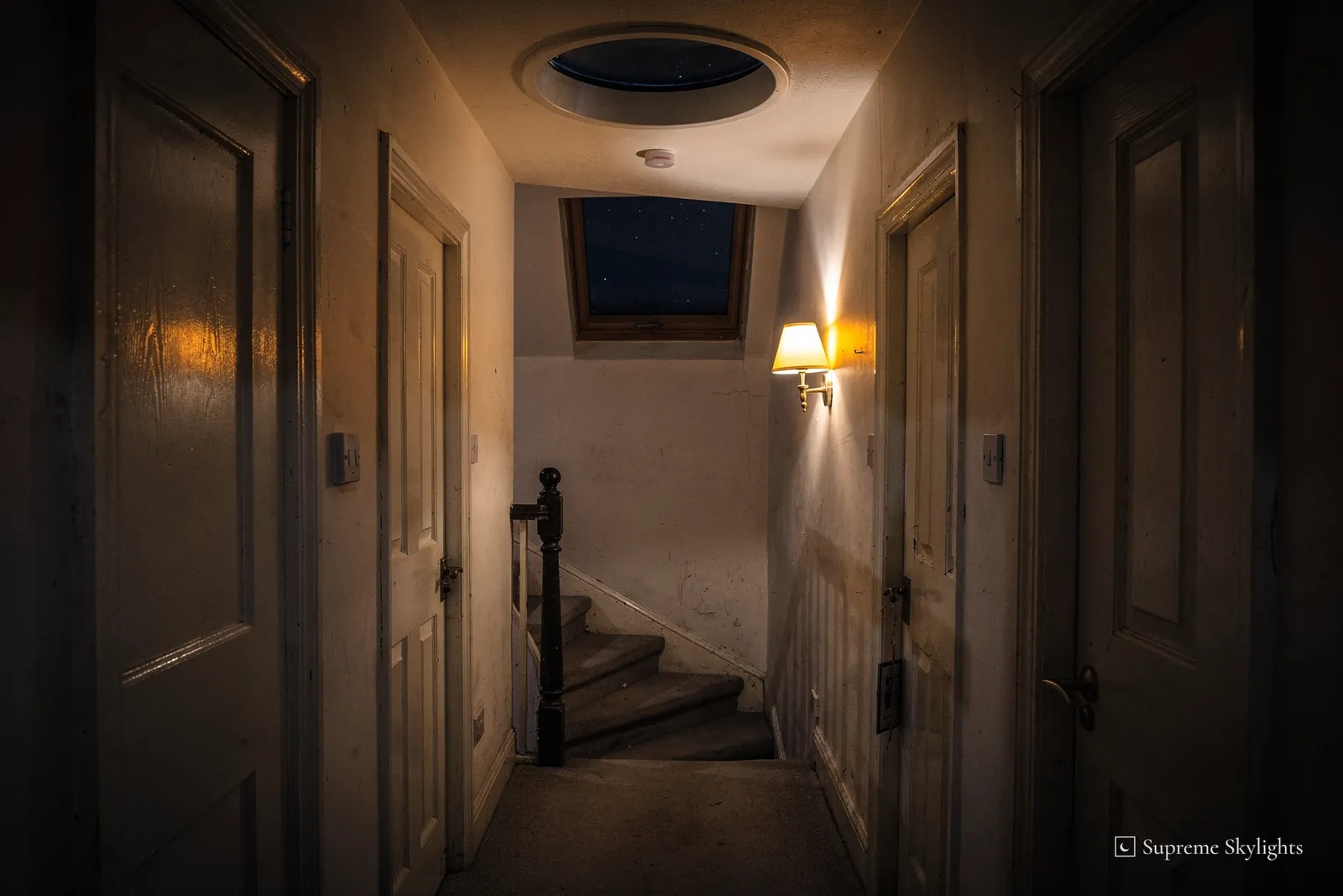 A narrow terraced-house landing at night, a round sun tunnel diffuser dark in the ceiling and a wall lamp lighting the stairs