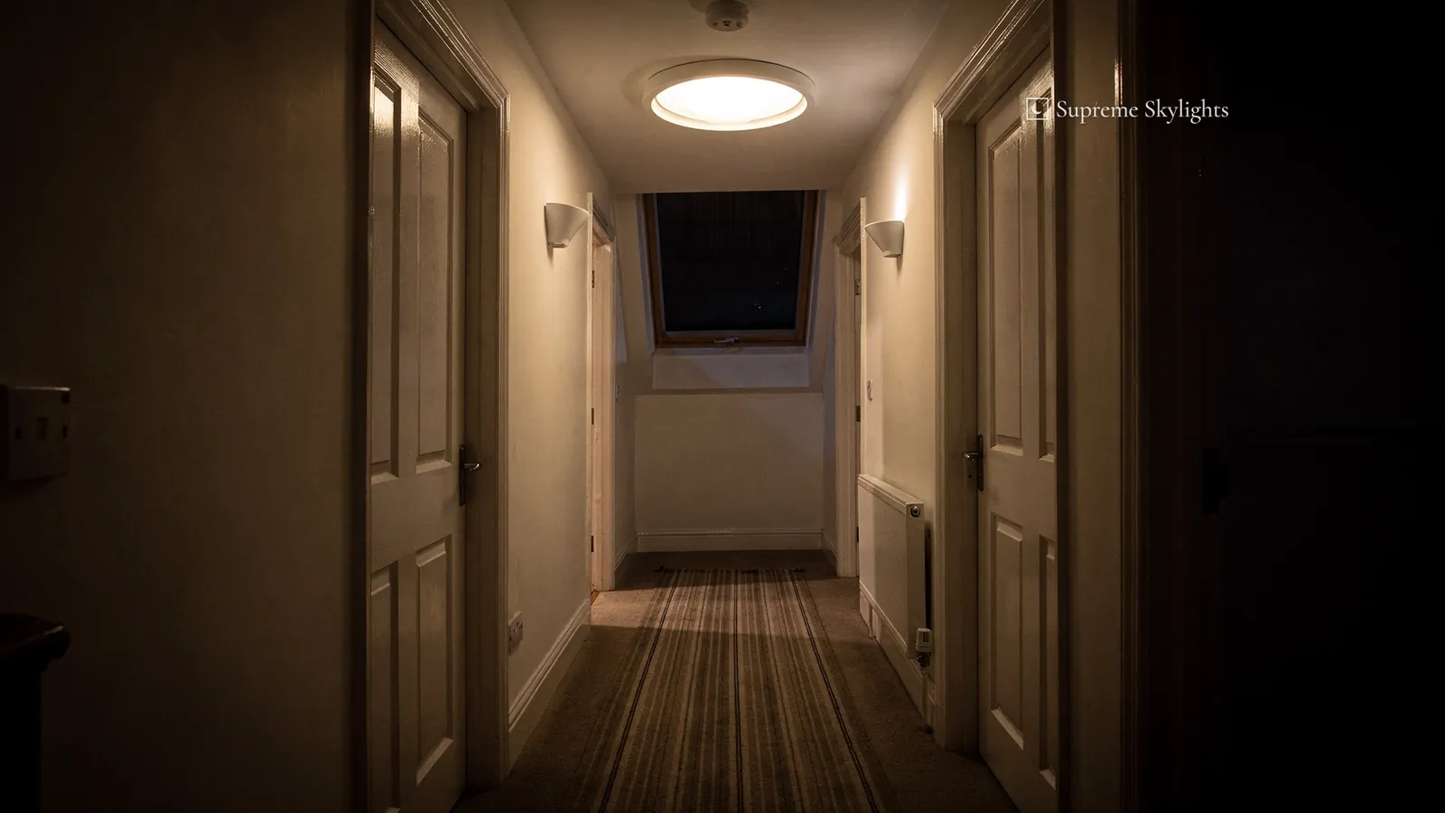 A narrow upstairs landing after dark, wall lights on and the round sun tunnel diffuser in the ceiling unlit